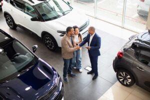 A mechanic explains car warranty details to a customer beside a car in a service garage.