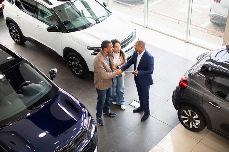 A mechanic explains car warranty details to a customer beside a car in a service garage.